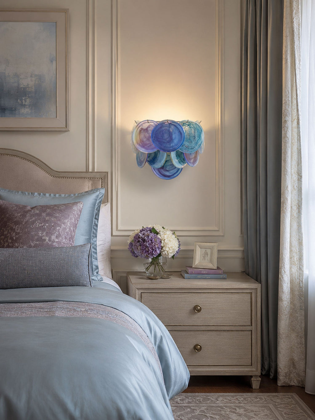 Small blue disc wall light from Clérachic glowing above a bedside table next to a bed dressed in blue and mauve linen.