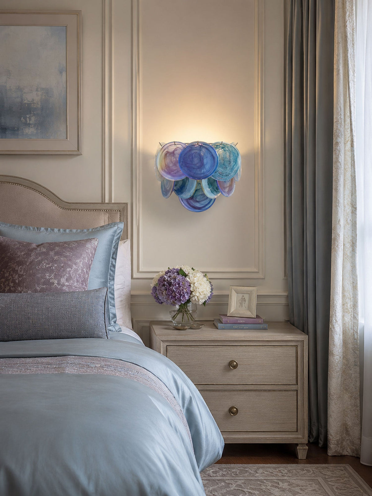 Small blue disc wall light from Clérachic glowing above a bedside table next to a bed dressed in blue and mauve linen.