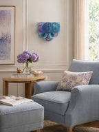 Blue and violet disc wall light from Clérachic above a side table with lilac flowers, next to a grey armchair by a window.