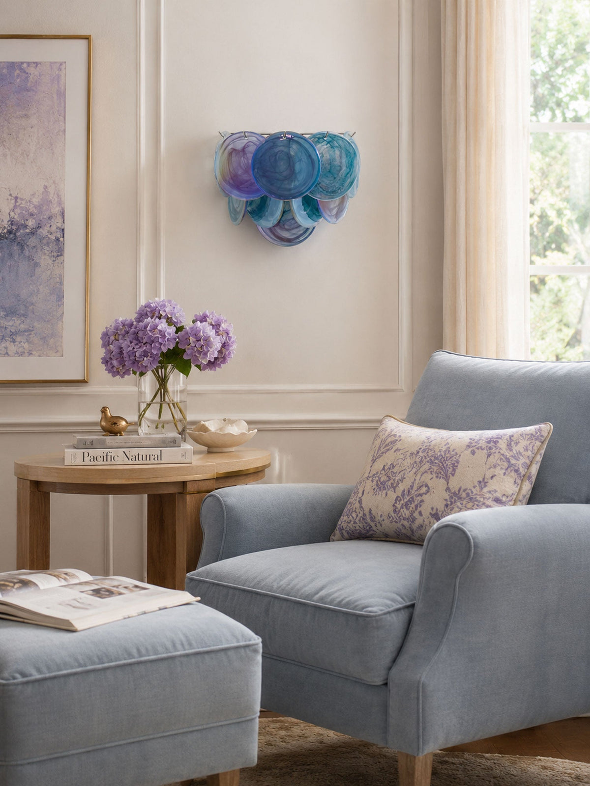 Blue and violet disc wall light from Clérachic above a side table with lilac flowers, next to a grey armchair by a window.