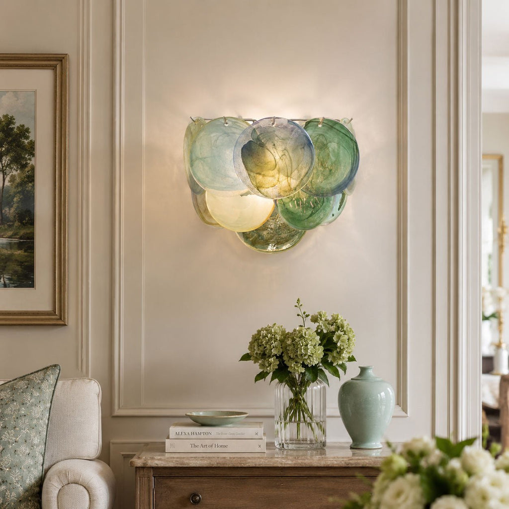 Close, lit view of a green and blue glass-disc wall light from Clérachic in a panelled room, with green hydrangeas on a console below.