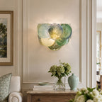 Close, lit view of a green and blue glass-disc wall light from Clérachic in a panelled room, with green hydrangeas on a console below.
