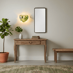 Small green glass-disc wall light from Clérachic glowing on a plain wall above a wooden console, with a tall mirror and a leafy plant.