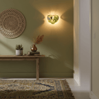 Olive-green glass-disc wall light from Clérachic lit on a sage wall, with a woven wall hanging and a wooden console beside it.