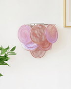 Pink and mauve glass-disc wall light from Clérachic with a chrome-coloured frame, mounted on a bright white wall beside a green leaf.