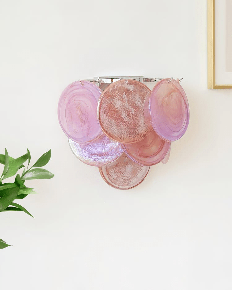 Pink and mauve glass-disc wall light from Clérachic with a chrome-coloured frame, mounted on a bright white wall beside a green leaf.