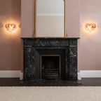 Two peach-toned disc wall lights from Clérachic glowing either side of a black marble fireplace and a gilt mirror on dusky pink walls.