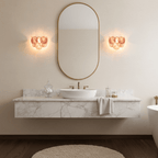 Pair of peach-pink disc wall lights from Clérachic either side of an oval gold-framed mirror over a marble floating vanity and a white basin.