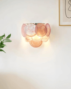 Lit pink and peach glass-disc wall light from Clérachic glowing on a white wall, with a leaf and a framed line drawing at the edges.