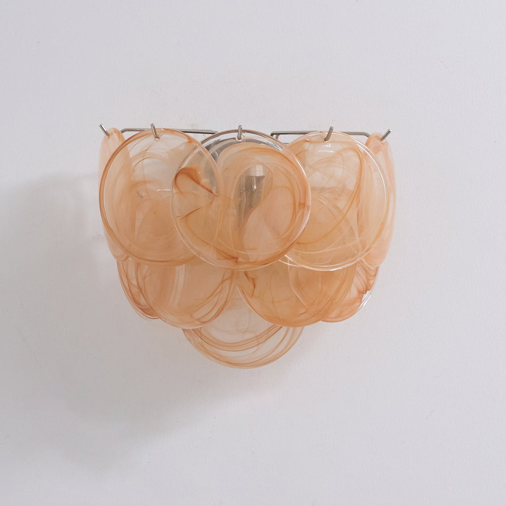 Unlit orange glass-disc wall light from Clérachic with swirled amber-peach discs layered in three tiers on a pale wall.