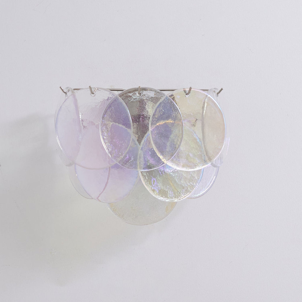 Unlit clear glass-disc wall light from Clérachic with iridescent discs showing a faint lilac sheen on a light grey wall.