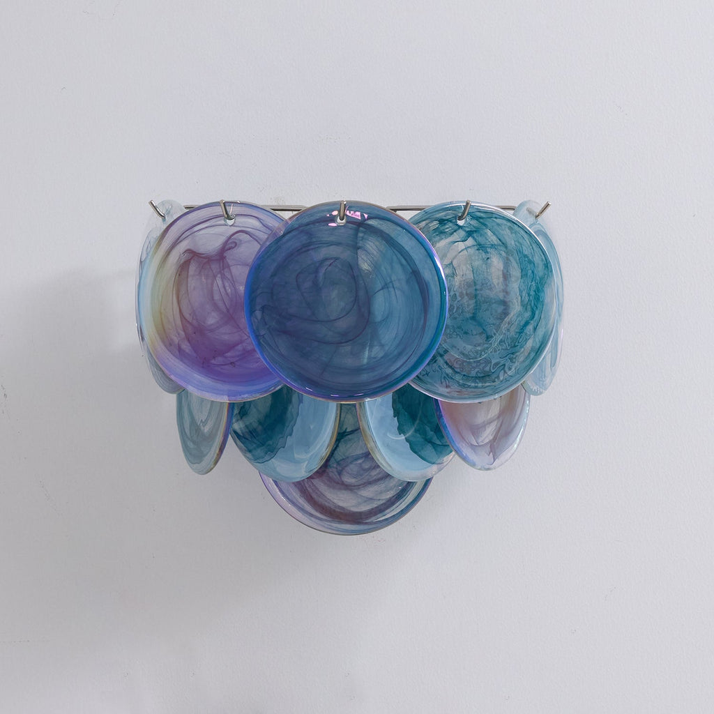 Unlit blue glass-disc wall light from Clérachic in teal, navy and violet swirls, shown straight on against a light wall.