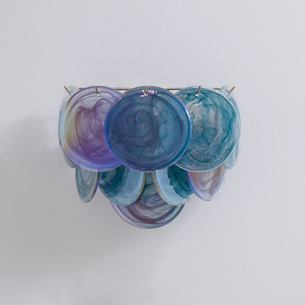 Unlit blue glass-disc wall light from Clérachic in teal, navy and violet swirls, shown straight on against a light wall.
