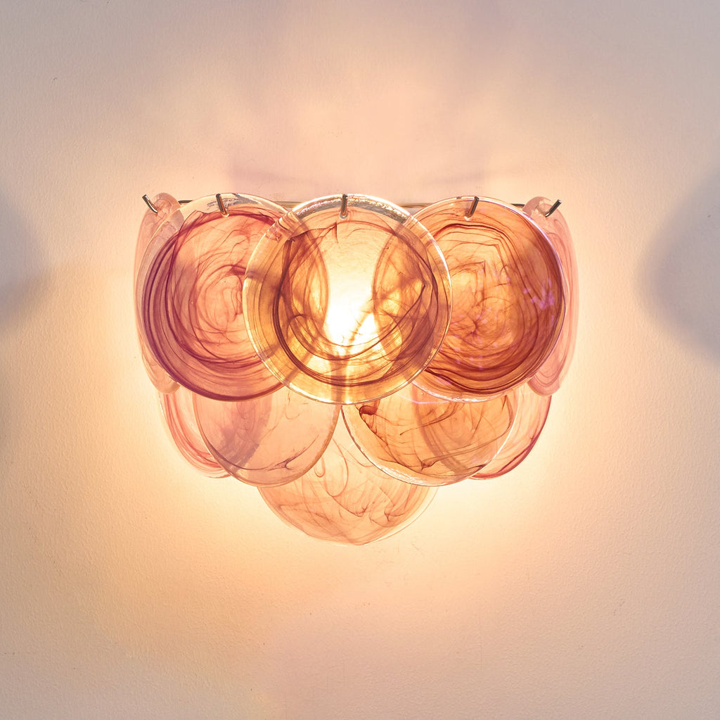 Orange glass-disc wall light from Clérachic lit, glowing warm with amber and rose swirls and a bright bulb behind the discs.