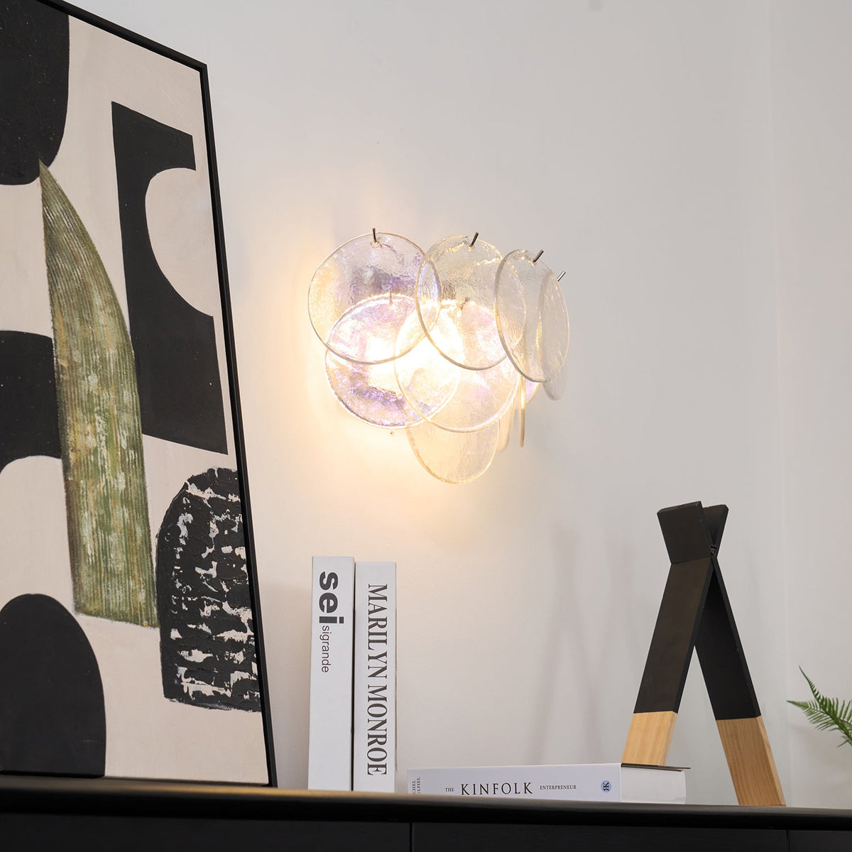 Clear iridescent disc wall light from Clérachic lit on a white wall beside a large abstract artwork and a wooden triangle ornament.