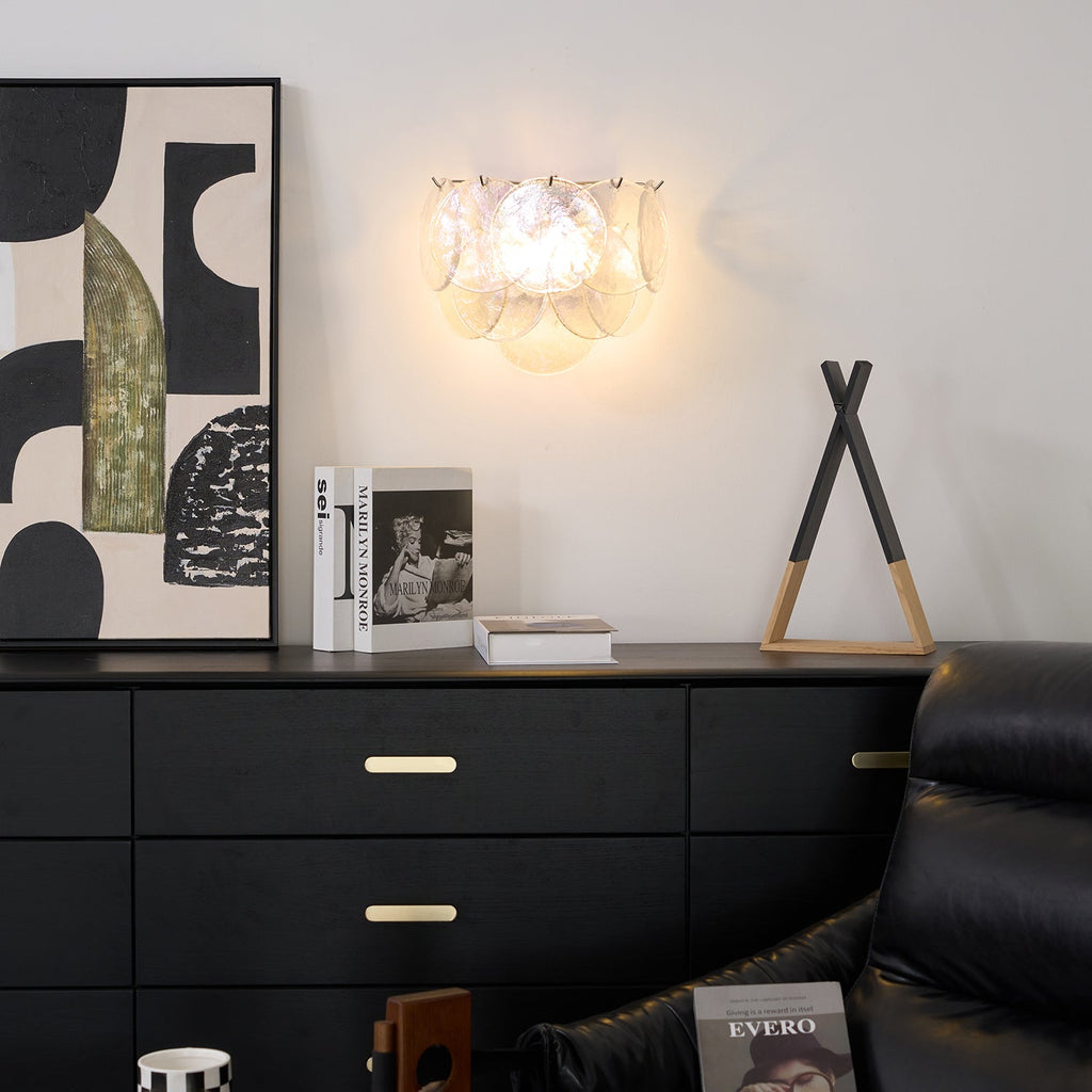 Lit iridescent glass-disc wall light from Clérachic above a black chest of drawers, with abstract art and a small ornament beside it.