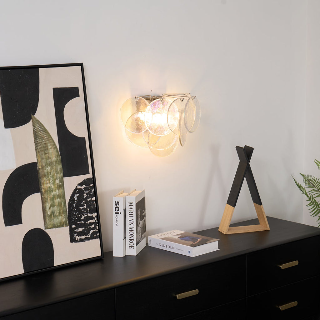 Clear glass-disc wall light from Clérachic lit above a black sideboard, with books and a wooden triangle ornament below.