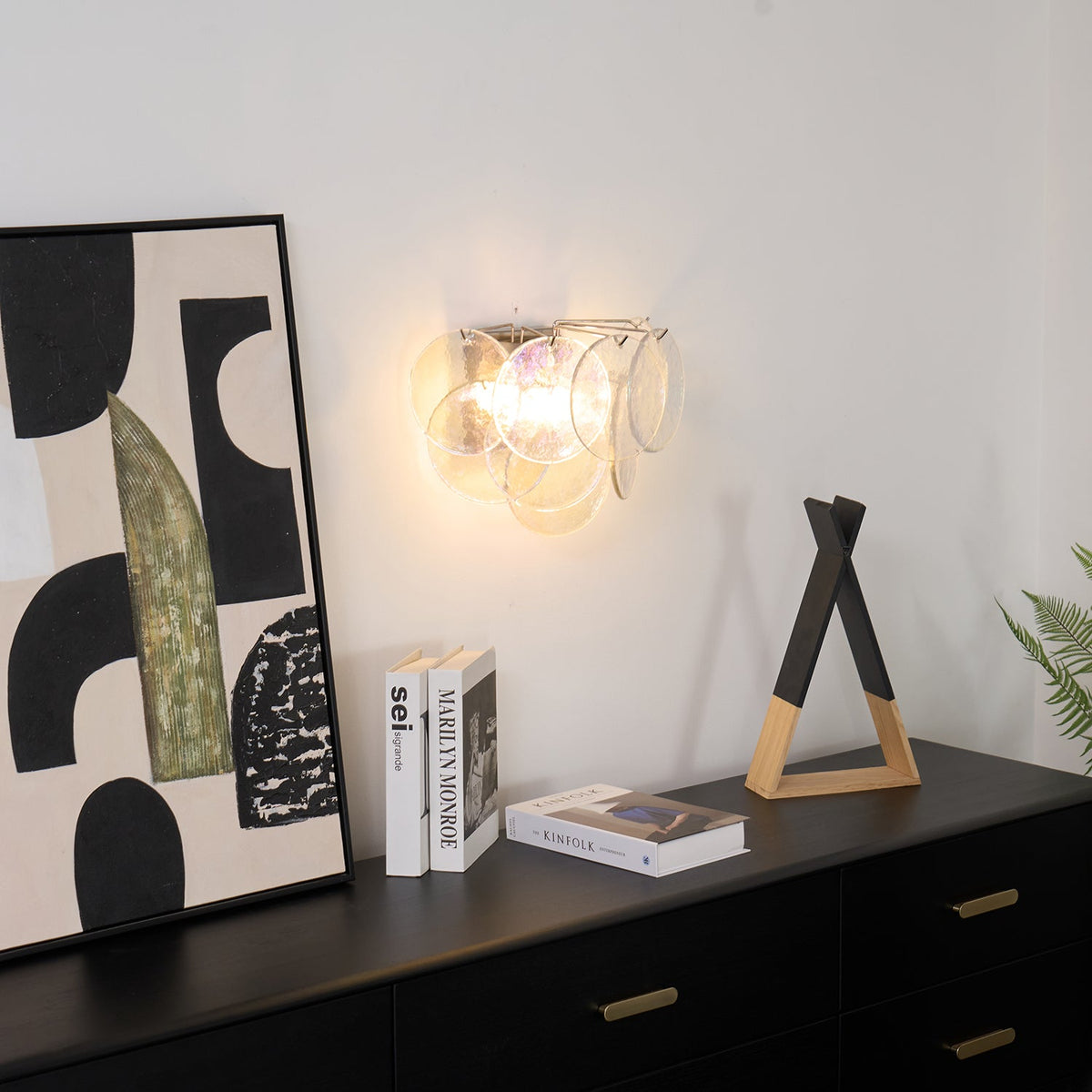 Clear glass-disc wall light from Clérachic lit above a black sideboard, with books and a wooden triangle ornament below.