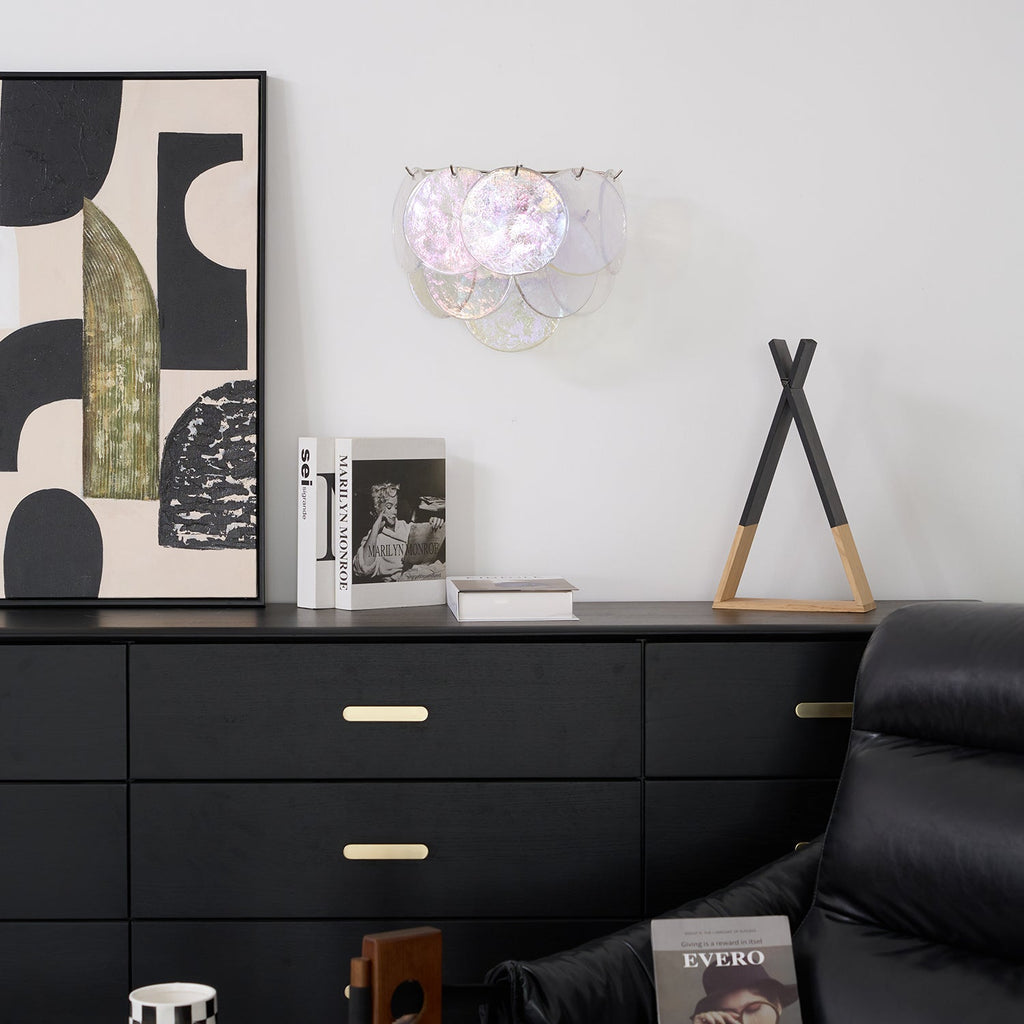 Unlit iridescent glass-disc wall light from Clérachic on a white wall above a black chest of drawers with brass handles.