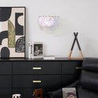 Unlit iridescent glass-disc wall light from Clérachic on a white wall above a black chest of drawers with brass handles.