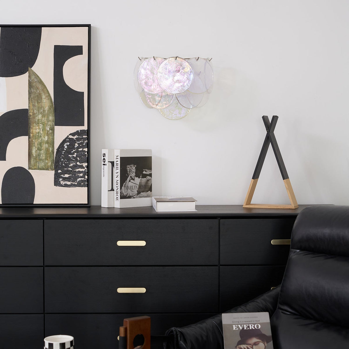 Unlit iridescent glass-disc wall light from Clérachic on a white wall above a black chest of drawers with brass handles.