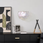 Wall light from Clérachic with pearly iridescent discs on a white wall, above a black sideboard with books and abstract art.