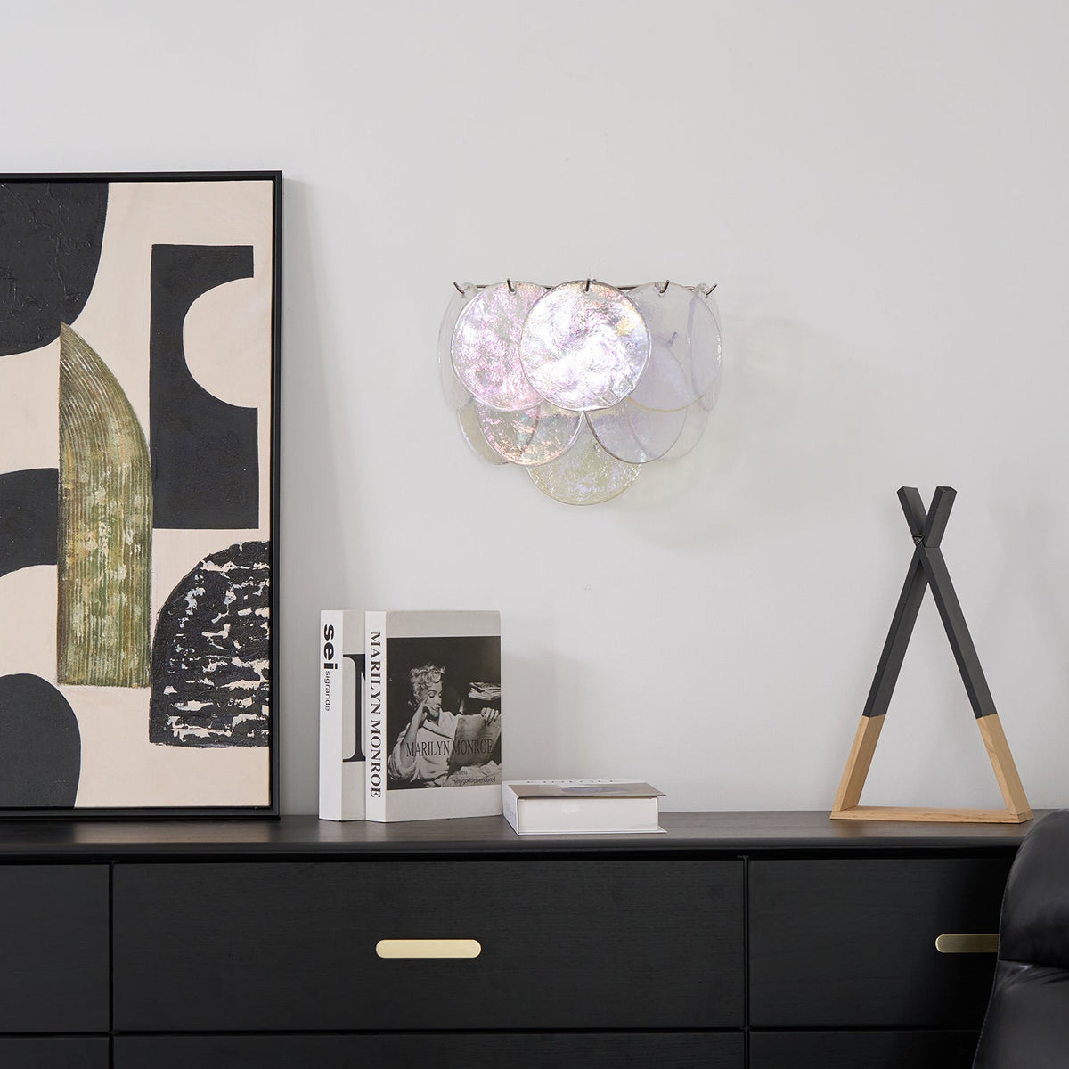 Wall light from Clérachic with pearly iridescent discs on a white wall, above a black sideboard with books and abstract art.