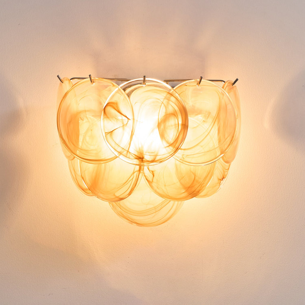 Close view of orange swirled glass discs on this Clérachic wall light, glowing amber with the bulb visible through the glass.