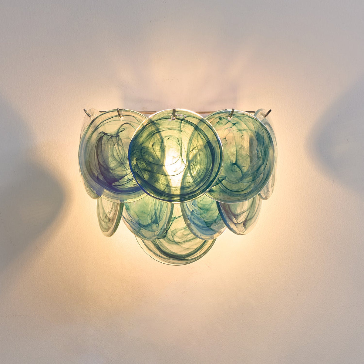 Close view of blue and teal swirled glass discs on this Clérachic wall light, lit warm against a pale wall.