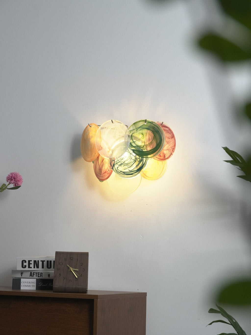 Small multicolour disc wall light from Clérachic glowing on a grey wall, with a desk clock, books and leaves in the foreground.