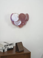 Unlit pink and lilac glass-disc wall light from Clérachic on a grey wall, above a wooden cabinet with books and a clock.