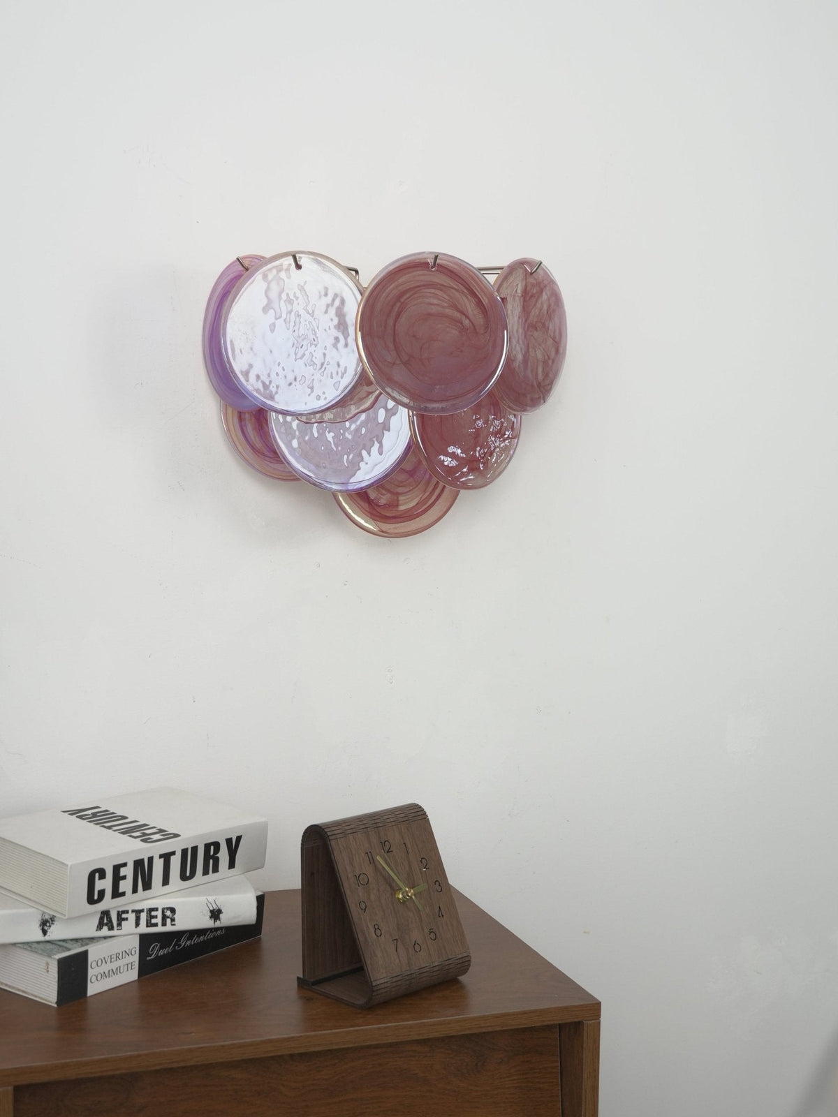 Unlit pink and lilac glass-disc wall light from Clérachic on a grey wall, above a wooden cabinet with books and a clock.