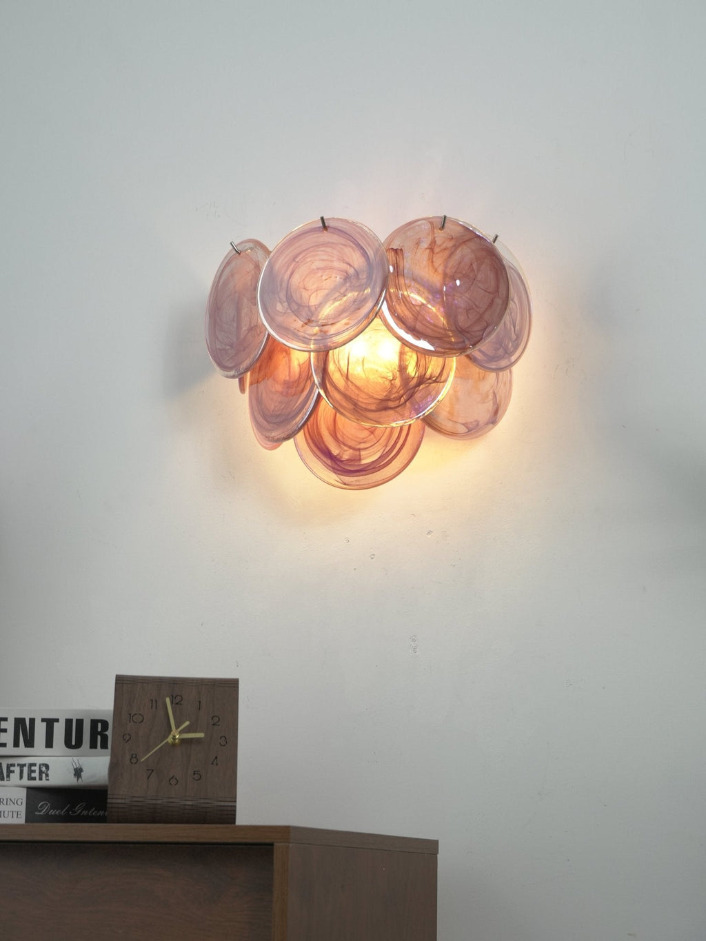 Lit pink and amber glass-disc wall light from Clérachic glowing on a grey wall above a wooden cabinet with a clock.