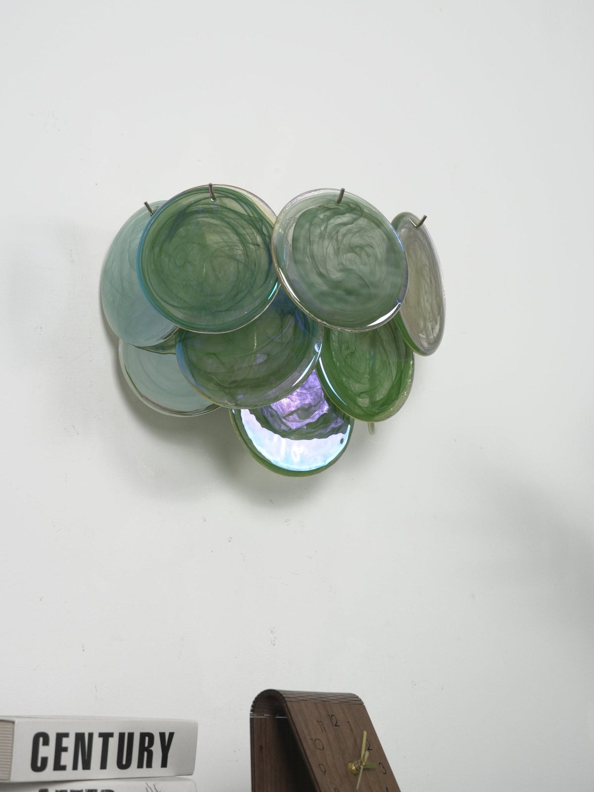 Unlit green glass-disc wall light from Clérachic with a single iridescent disc at the base, above a stack of books and a clock.