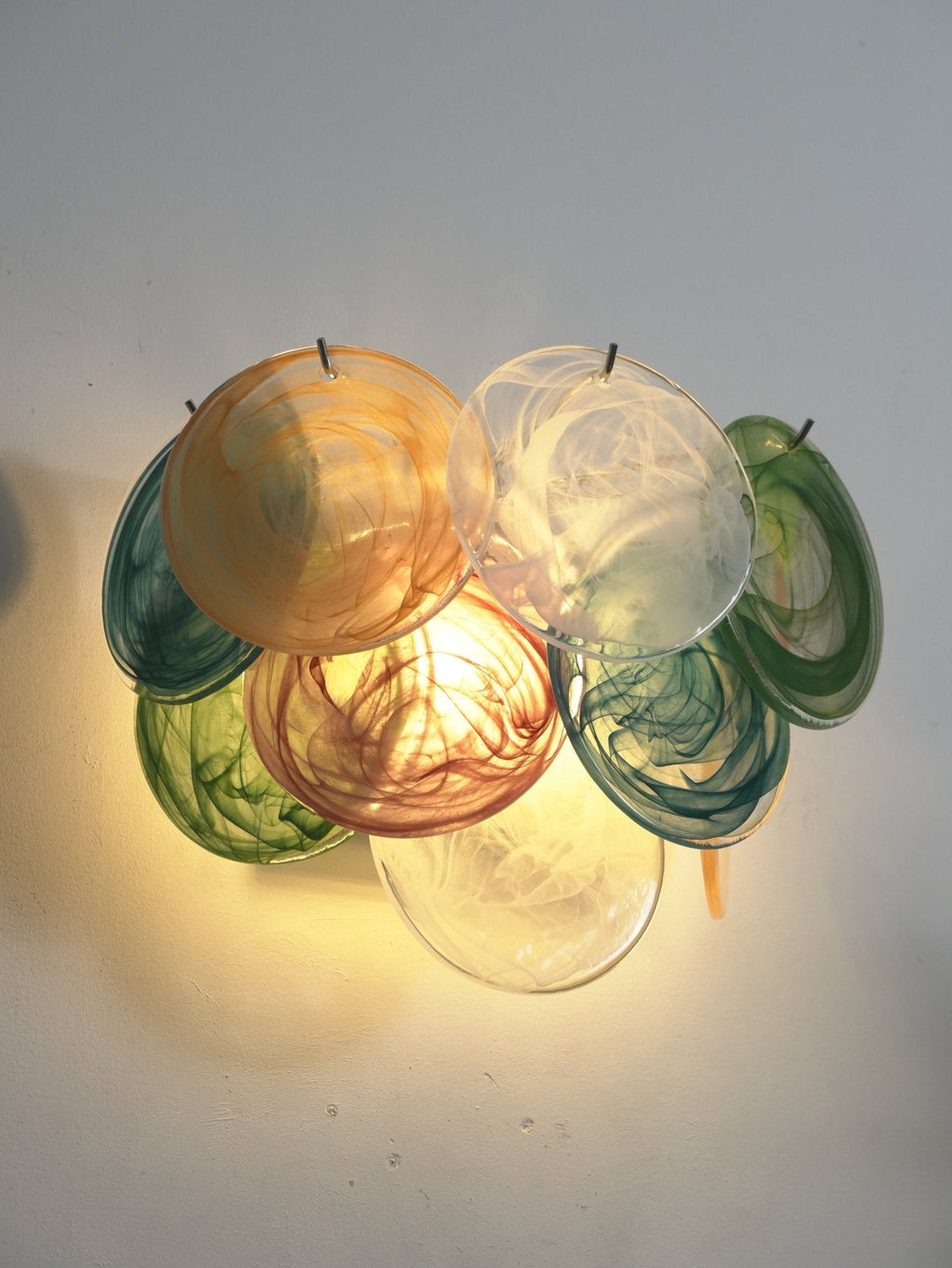 Lit multicolour wall light from Clérachic with orange, white, teal, rose and green glass discs glowing on a pale grey wall.