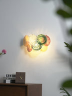 Small lit multicolour wall light from Clérachic on a grey wall, with a pink flower, a plant and a clock in the frame.