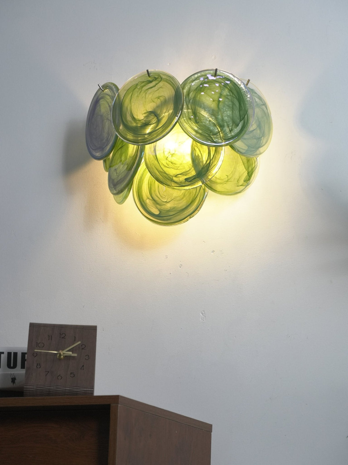 Lit green glass-disc wall light from Clérachic glowing yellow-green on a white wall above a wooden cabinet with a desk clock.
