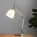 Lit white fabric shade on a silver jointed arm, standing on a wooden desk against a grey wall beside a leafy plant, from Clérachic.