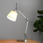 Lit white fabric shade on a silver jointed arm, standing on a wooden desk against a grey wall beside a leafy plant, from Clérachic.