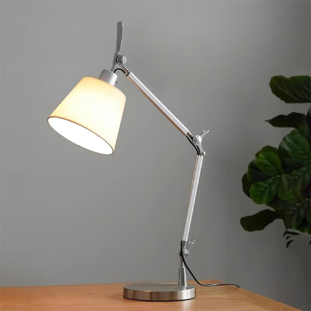 Lit white fabric shade on a silver jointed arm, standing on a wooden desk against a grey wall beside a leafy plant, from Clérachic.