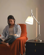 This Clérachic table lamp lights a woman reading in an orange armchair, with the silver arm reaching over a dark nightstand.