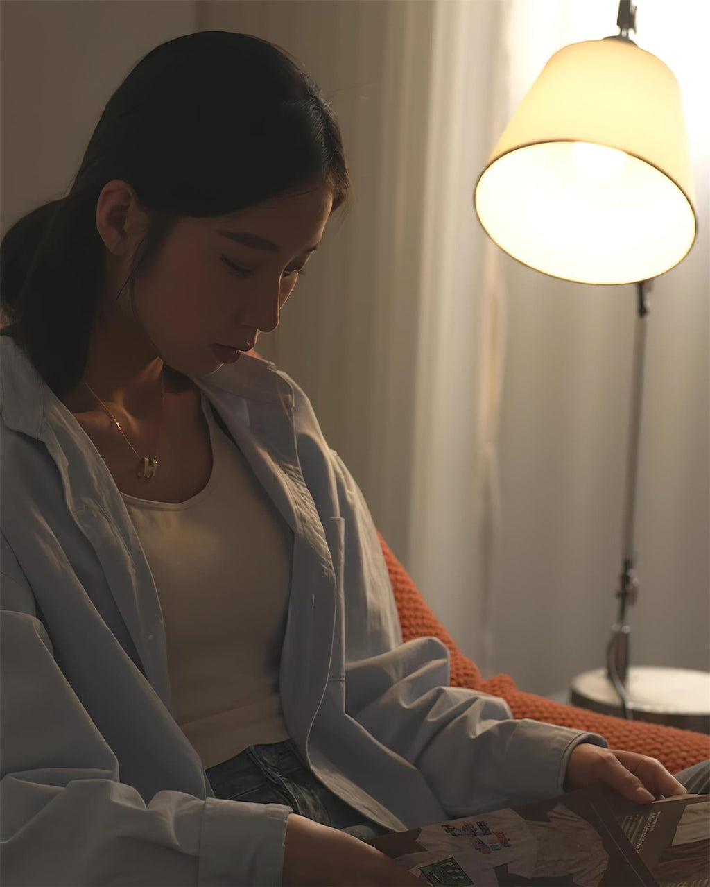 Soft light from the white shade of a Clérachic lamp in a dim room, with a woman in a pale shirt leafing through a magazine.
