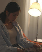 Soft light from the white shade of a Clérachic lamp in a dim room, with a woman in a pale shirt leafing through a magazine.