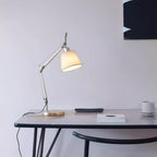 Silver arm lamp from Clérachic on a slim black desk with a coffee cup and a book, its white shade lit against a pale wall.