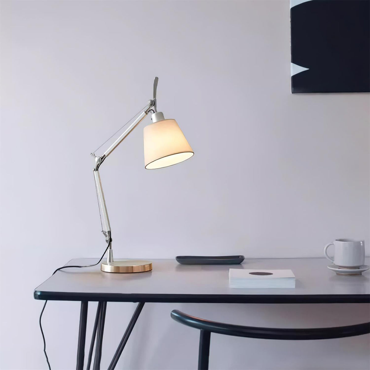 Silver arm lamp from Clérachic on a slim black desk with a coffee cup and a book, its white shade lit against a pale wall.