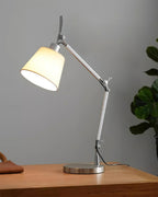 Side view of this Clérachic lamp on a wooden tabletop, the silver arm angled over a round metal base next to a green plant.