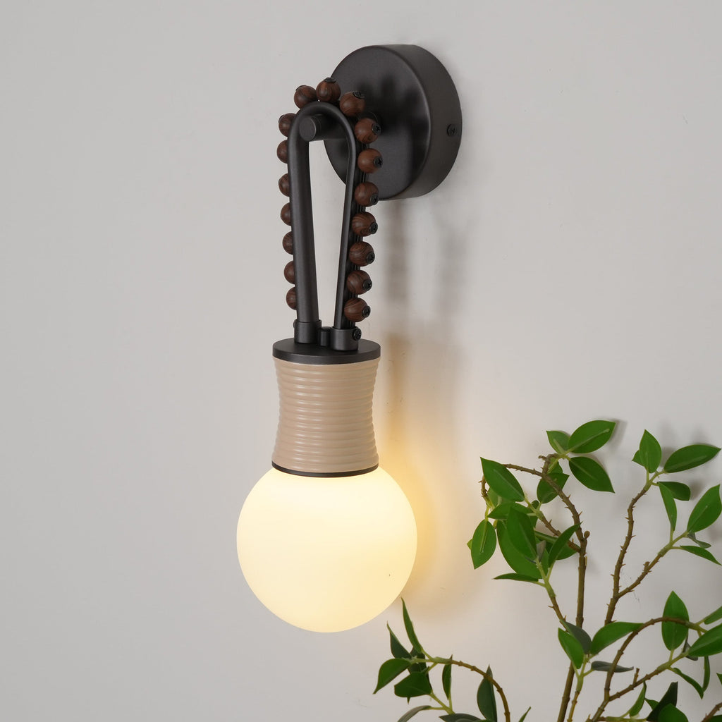 Clérachic Sytala sconce lit warm on a pale wall beside trailing greenery, opal globe glowing softly.