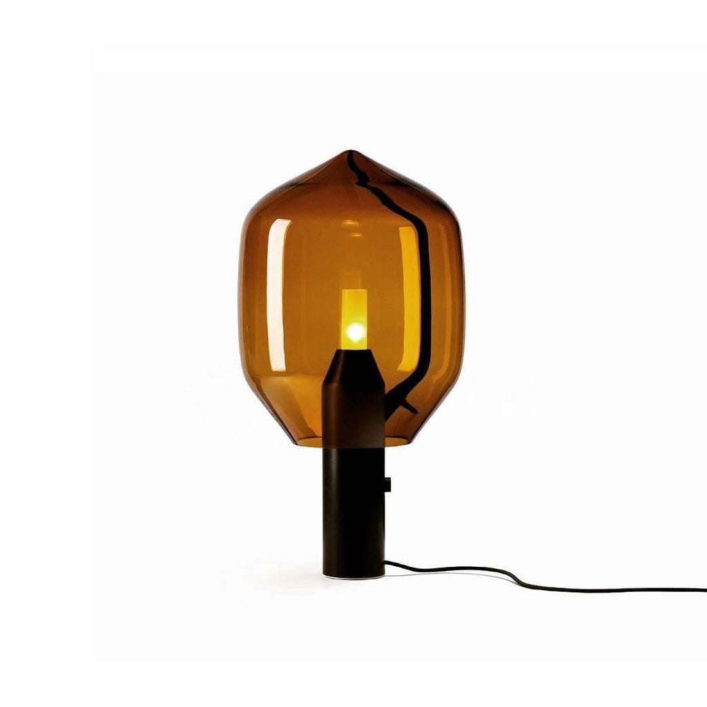 An amber-glass Clérachic table lamp on its black base, its inline switch and cord visible against a plain white backdrop.