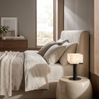 This black Clérachic table lamp glows softly on a cream pedestal nightstand beside an upholstered bed.