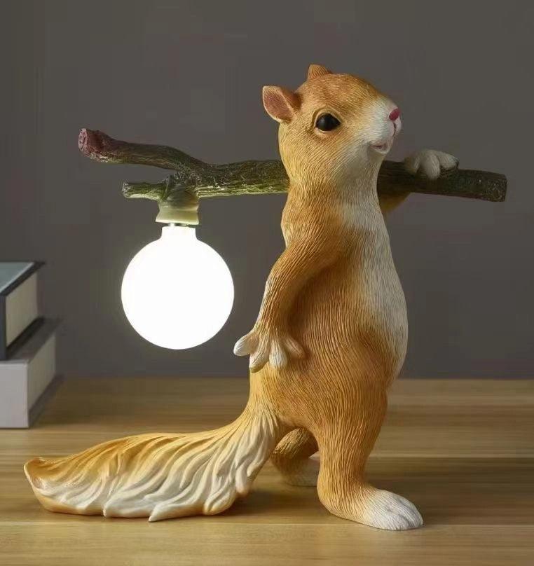 Clérachic squirrel lamp in a tan-gold finish, its round bulb glowing white, on a wooden shelf beside a stack of books.